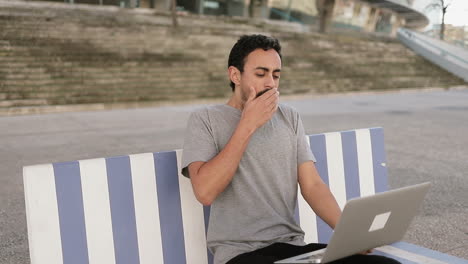 Tired-young-Arabic-man-with-dark-curly-hair-and-beard-in-grey-T-shirt-sitting-on-quay-on-striped-bench